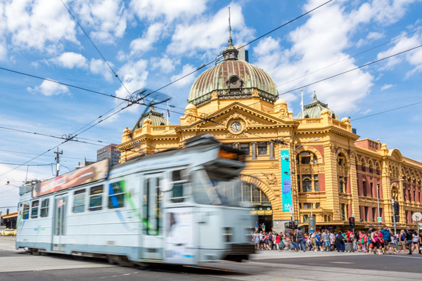Flinders street station and tram in Melbourne, Australia on a nice sunny day. The tram is moving and people are walking at the busy intersection in the background.